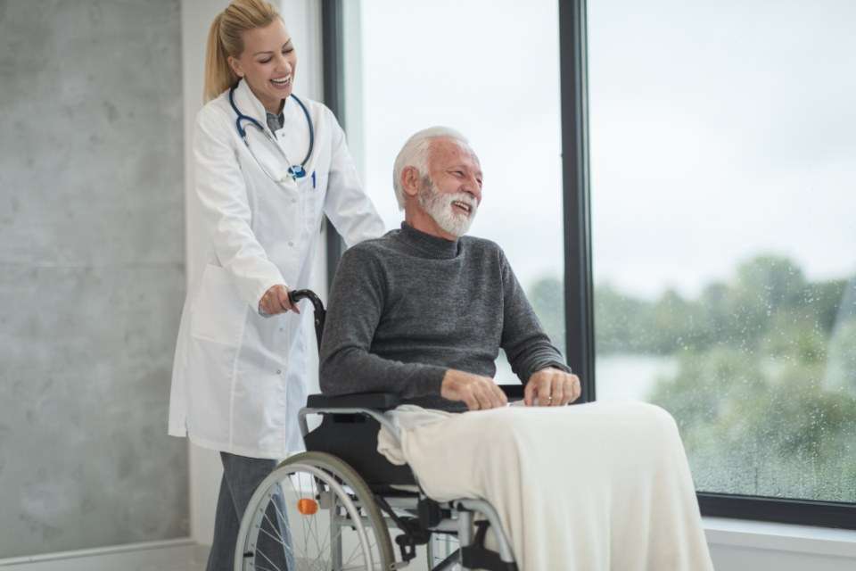 A doctor and an older man in a wheelchair share a laugh by a window.