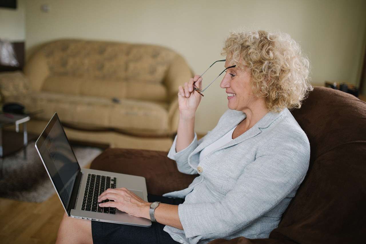 An older woman at home smiles at her laptop as she puts on her glasses.